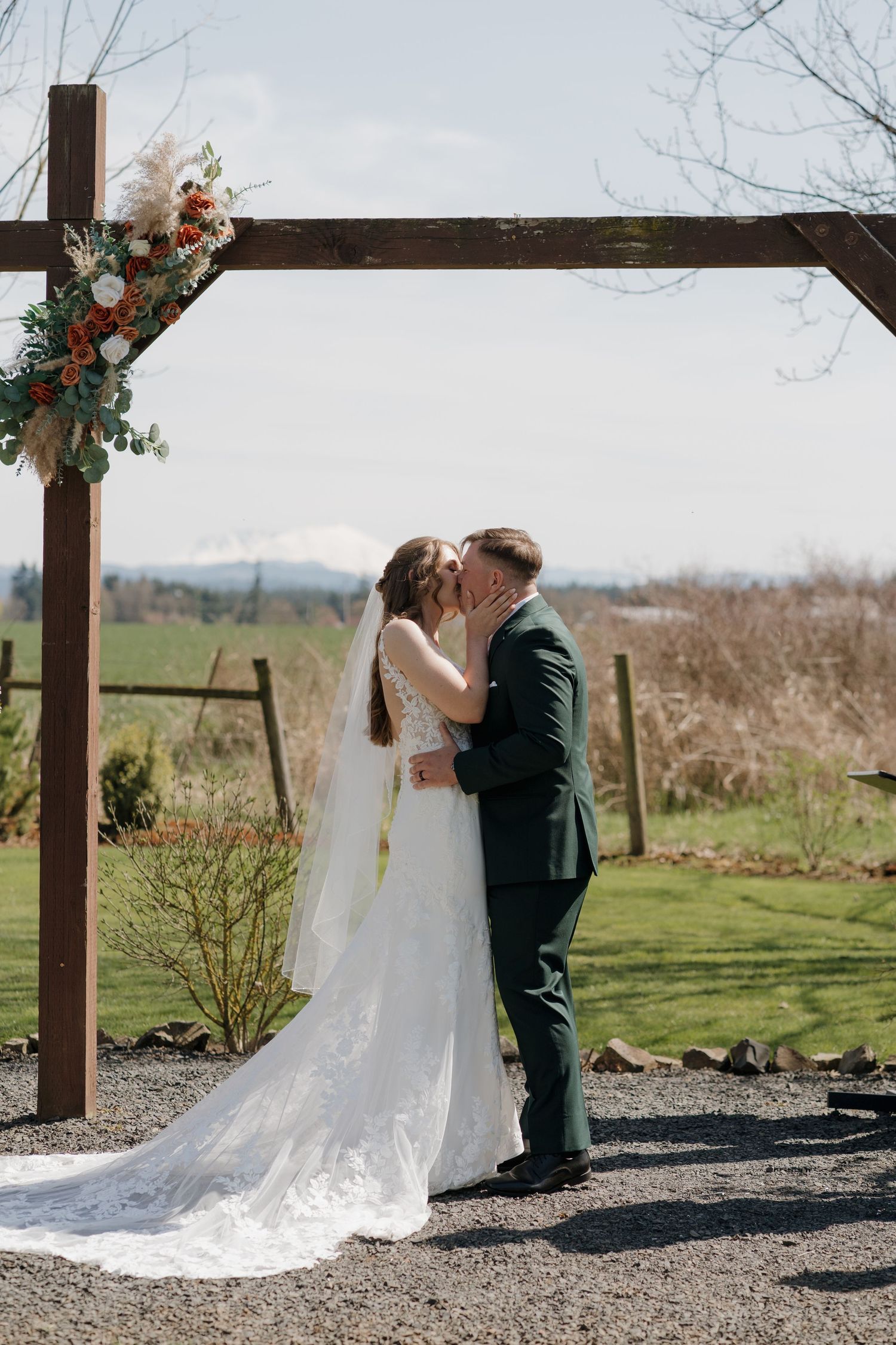 A couple shares a romantic moment on a large driftwood log against a backdrop of evergreen mountains and cloudy skies.
