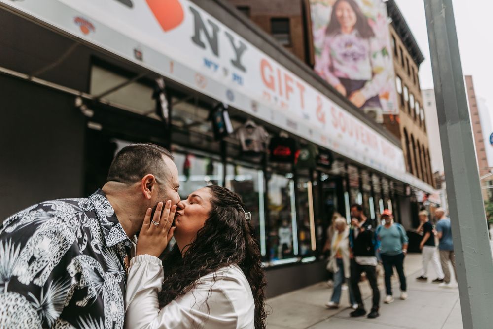 A couple shares a romantic kiss on a city street with storefronts and pedestrians in the background.