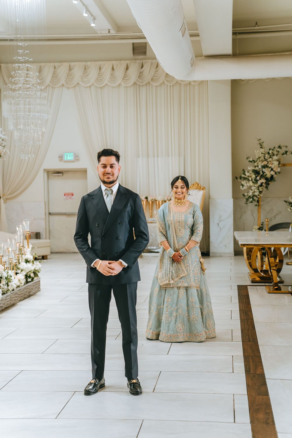 Couple in wedding attire pose in an elegantly decorated venue with white draping and floral arrangements.