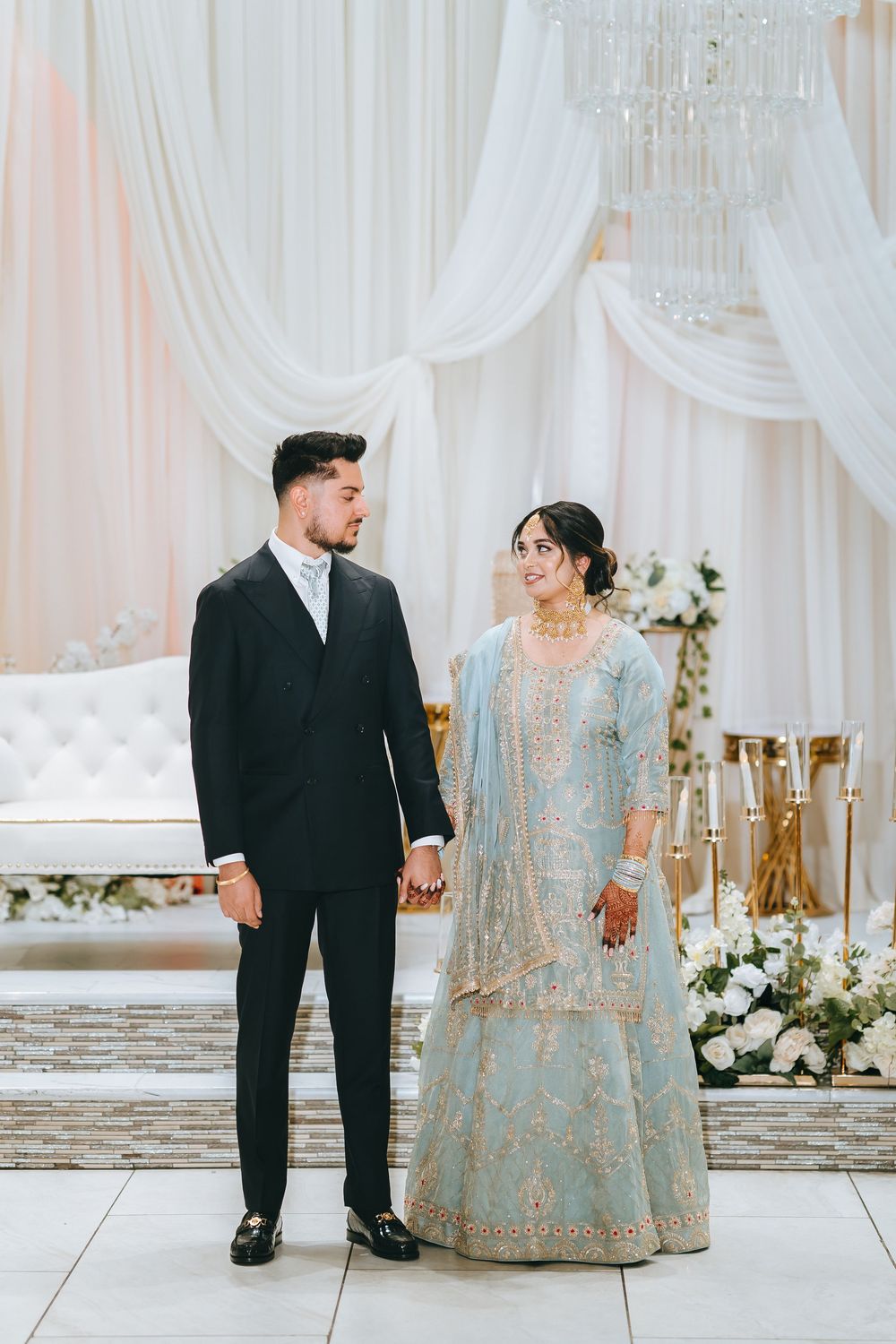A couple in traditional wedding attire share tender moments in an elegantly decorated white venue.