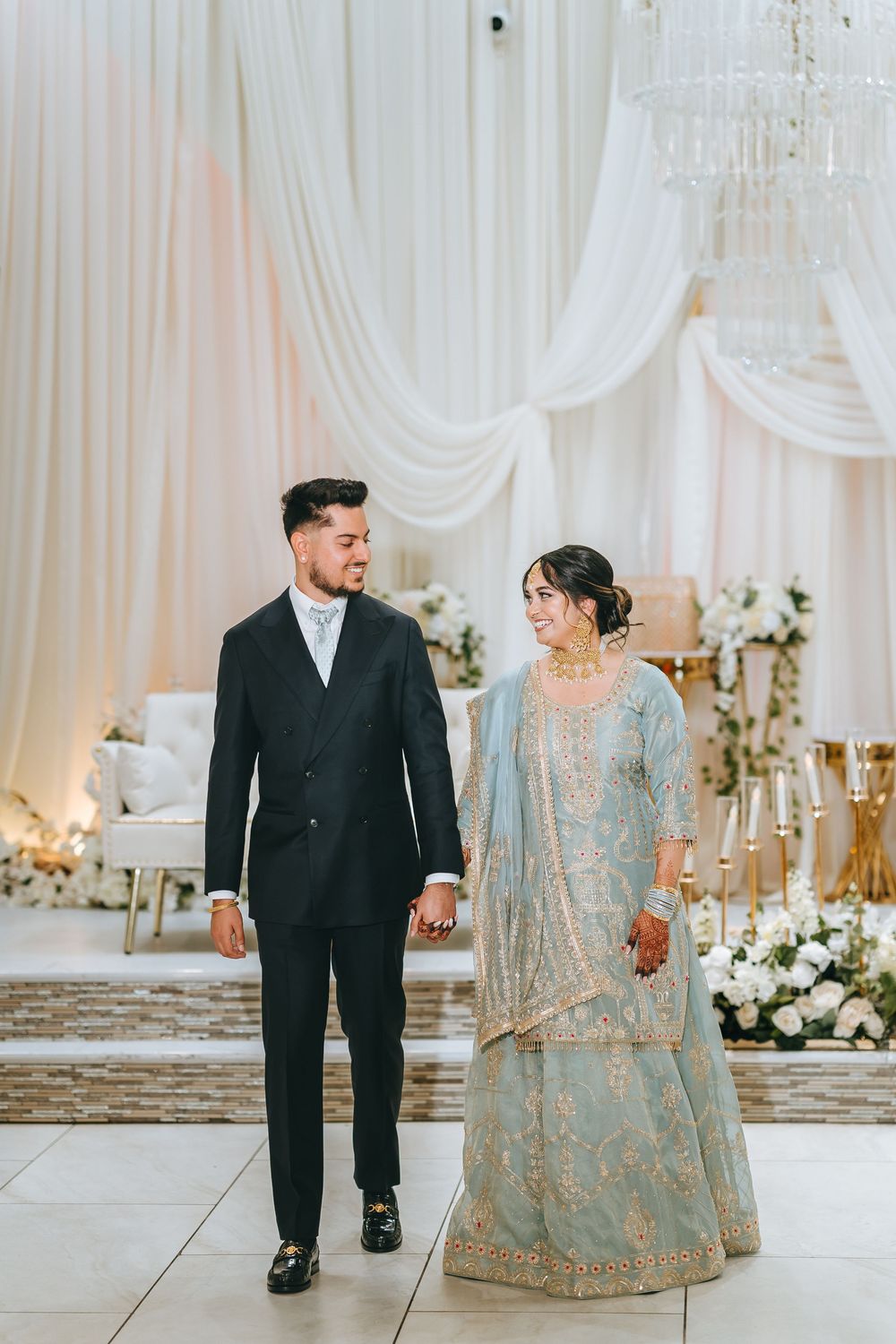 A couple in wedding attire stand together at an elegant reception with white drapes and floral decor.