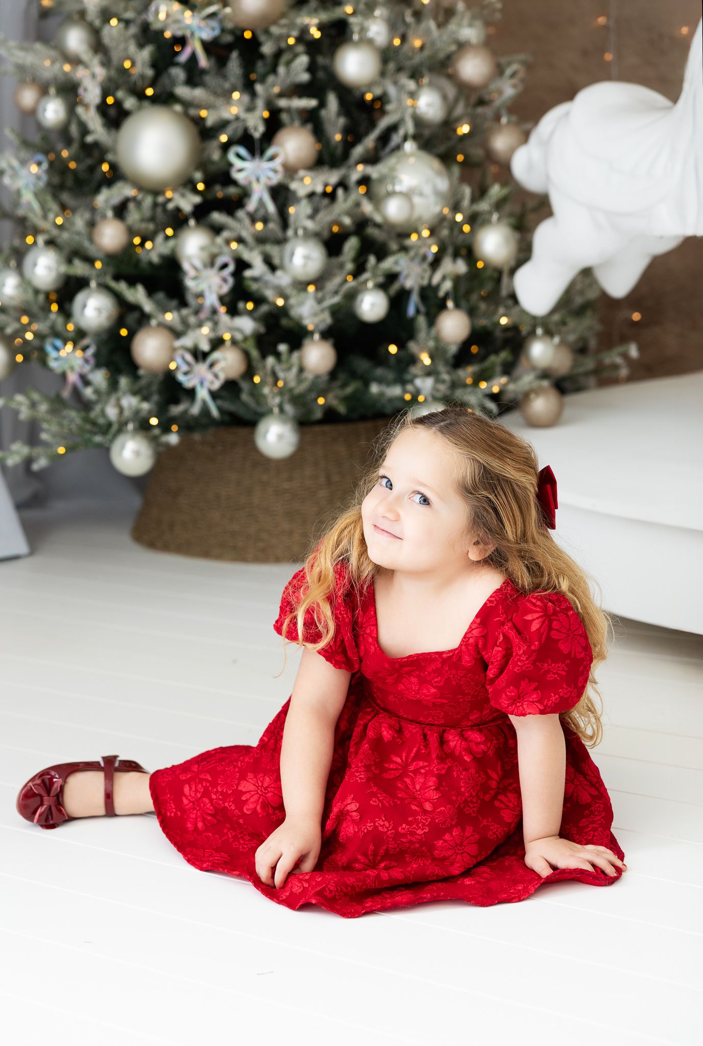 A child in a red velvet dress lays on a white floor with a decorated Christmas tree in the background.
