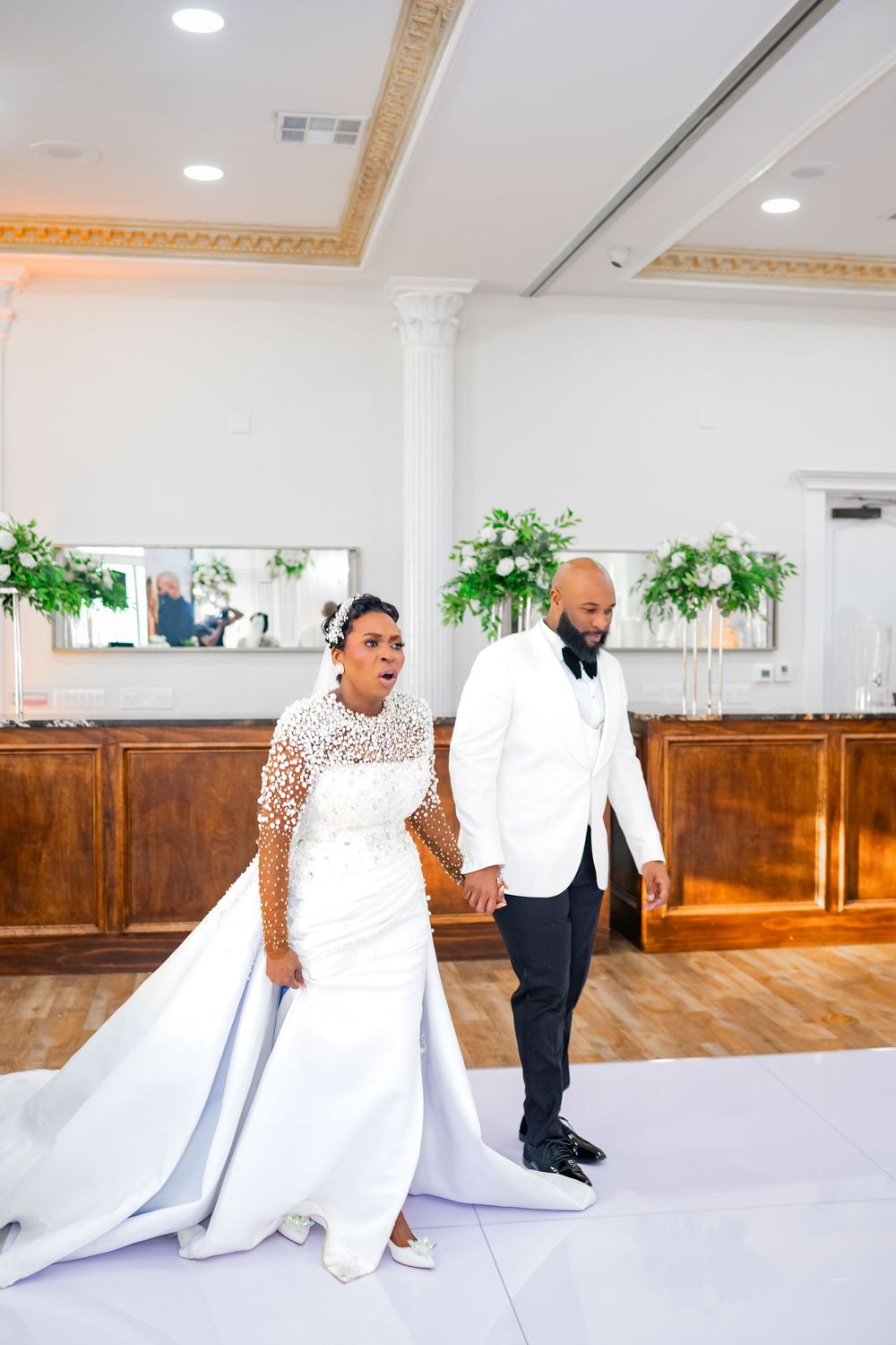Two people in formal attire set up white linens on a table in an elegant banquet hall with ornate crown molding.