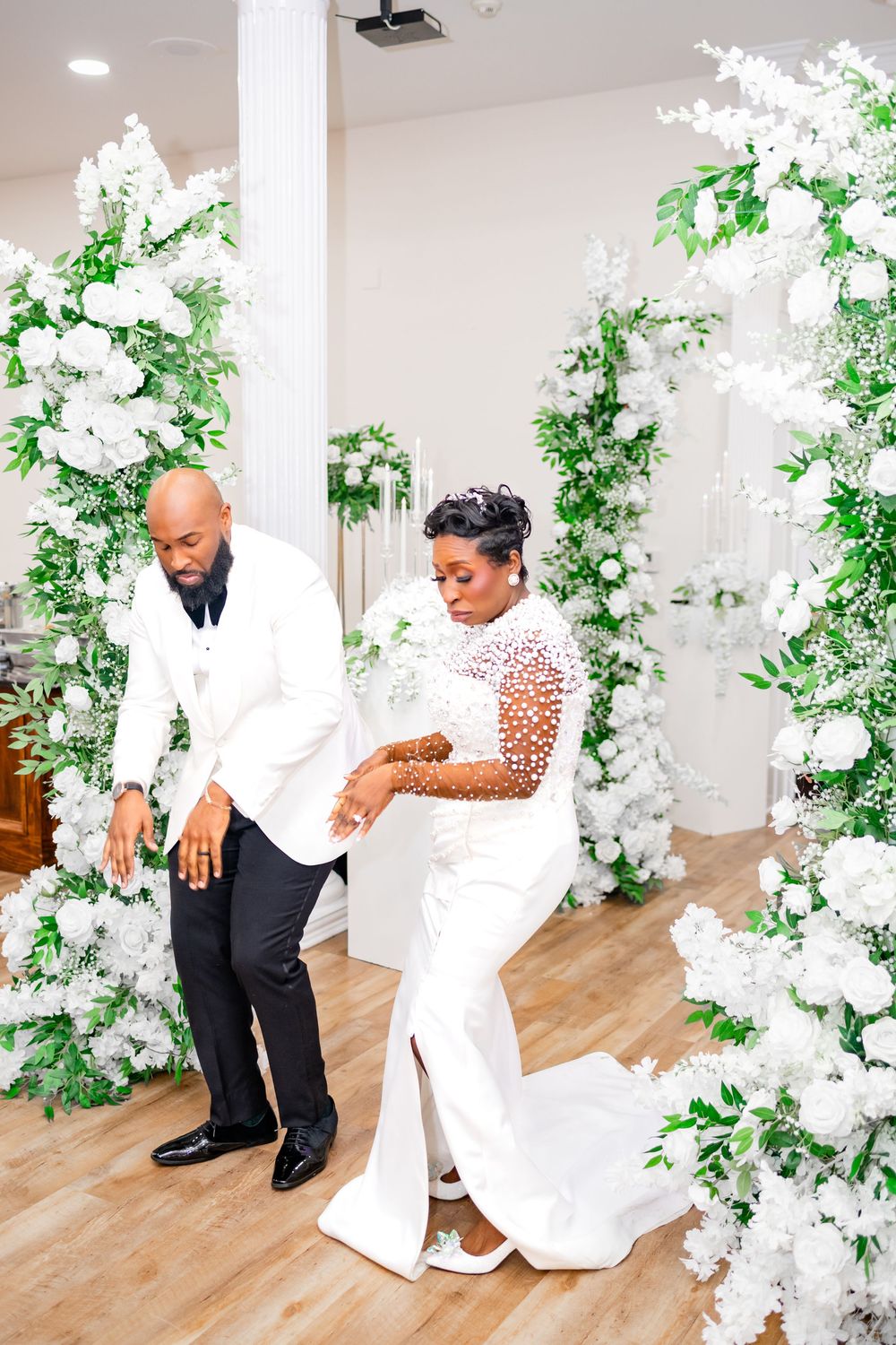 A couple shares a romantic dance moment surrounded by white floral arrangements and greenery in an elegant indoor setting.