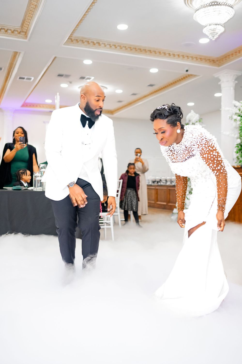 Elegant wedding reception scene showing couples dancing and celebrating in a bright white ballroom with ornate decor.