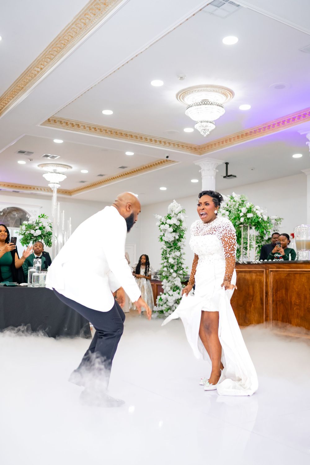 Dancing couple performs energetic moves on the dance floor at an elegant reception venue with ornate ceiling details.