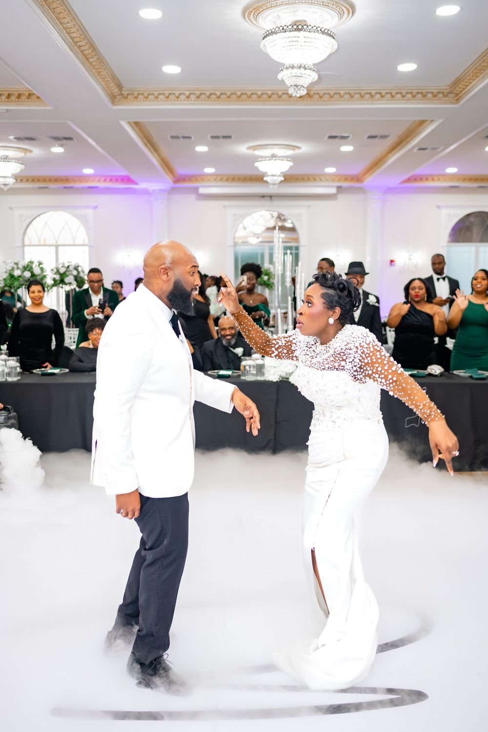 Dancers in elegant white attire move gracefully across an ornate ballroom floor under crystal chandeliers.