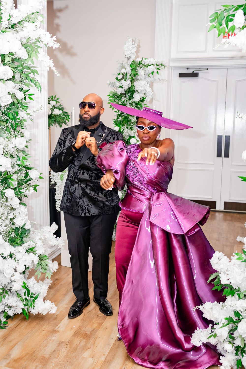 A couple poses playfully in formal attire, with one wearing a bright pink gown and another in black at an elegant venue.