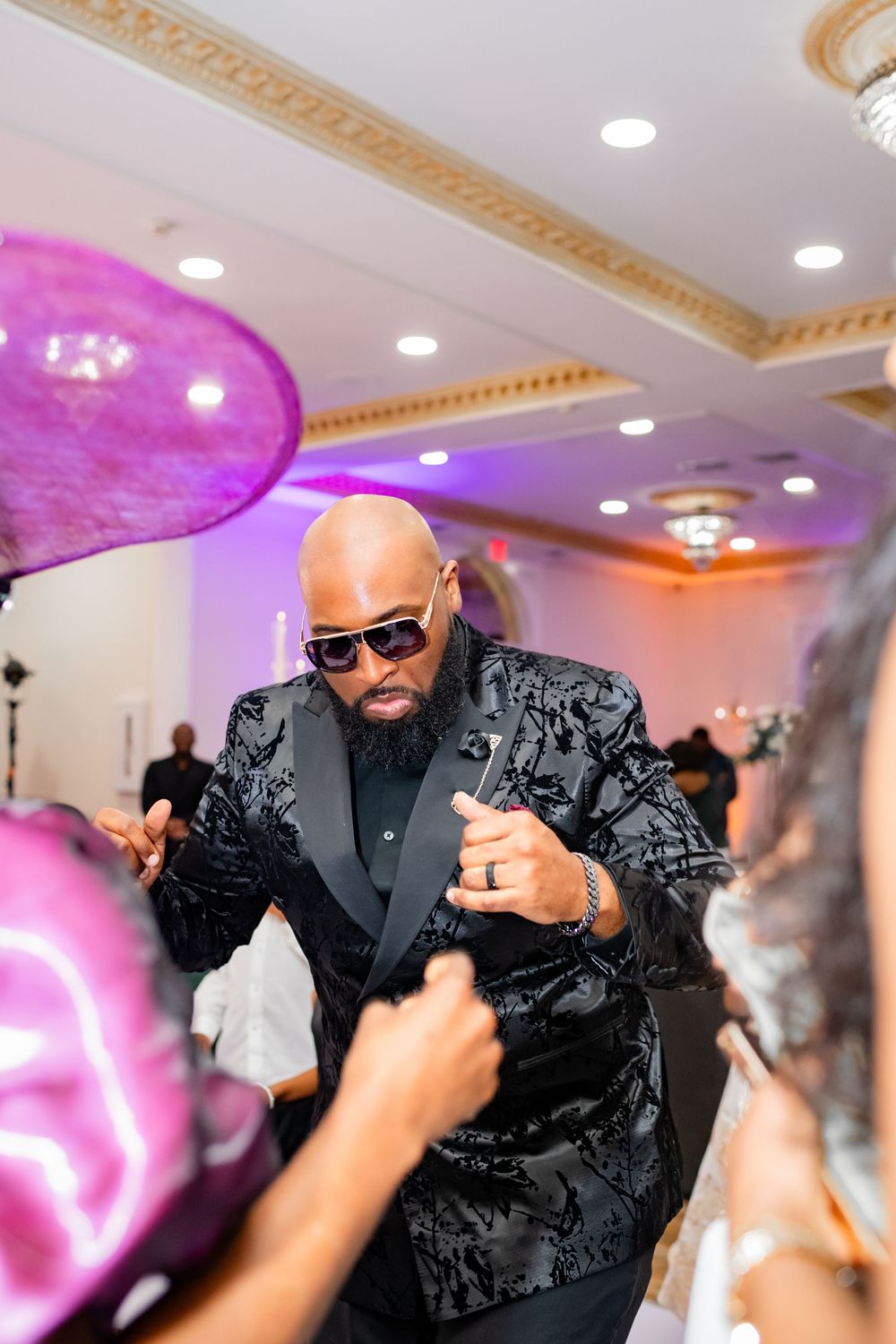 Wedding guests dance and celebrate with a man in a black patterned suit and sunglasses at an elegant reception venue.