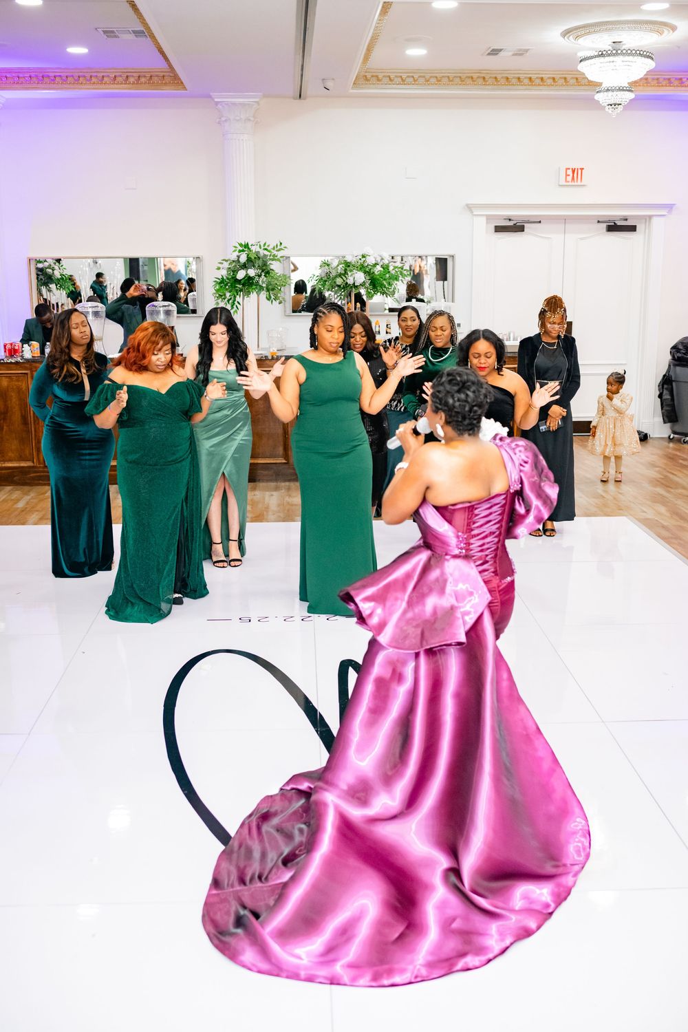 Wedding party members in green dresses dance with someone wearing a flowing pink gown in a brightly lit ballroom.