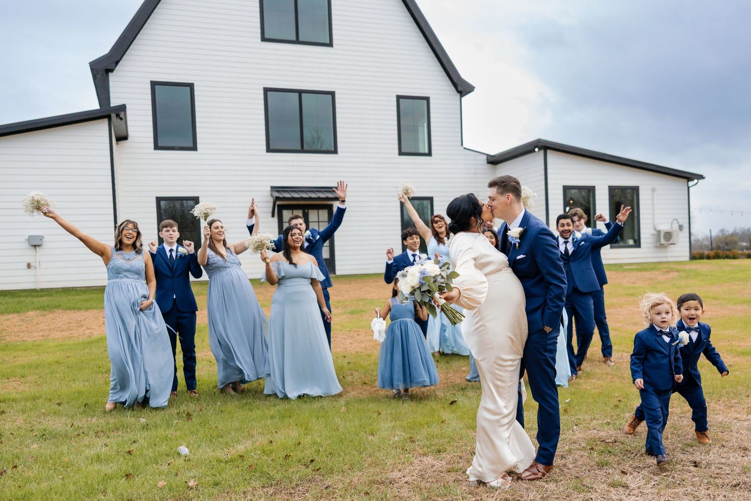 Wedding party in blue dresses and navy suits celebrates outside white barn venue.