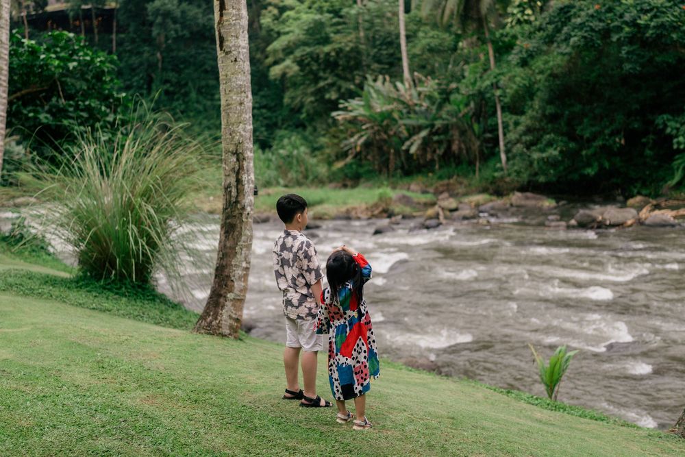 People gather for photos near a flowing river with trees and rocks in a natural outdoor setting.
