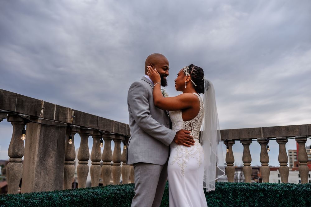 A couple in formal attire embraces on a balcony with stone columns under a cloudy sky.