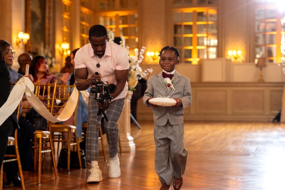 A young ring bearer in a gray suit walks down the aisle at an elegant wedding reception venue with glowing lights.