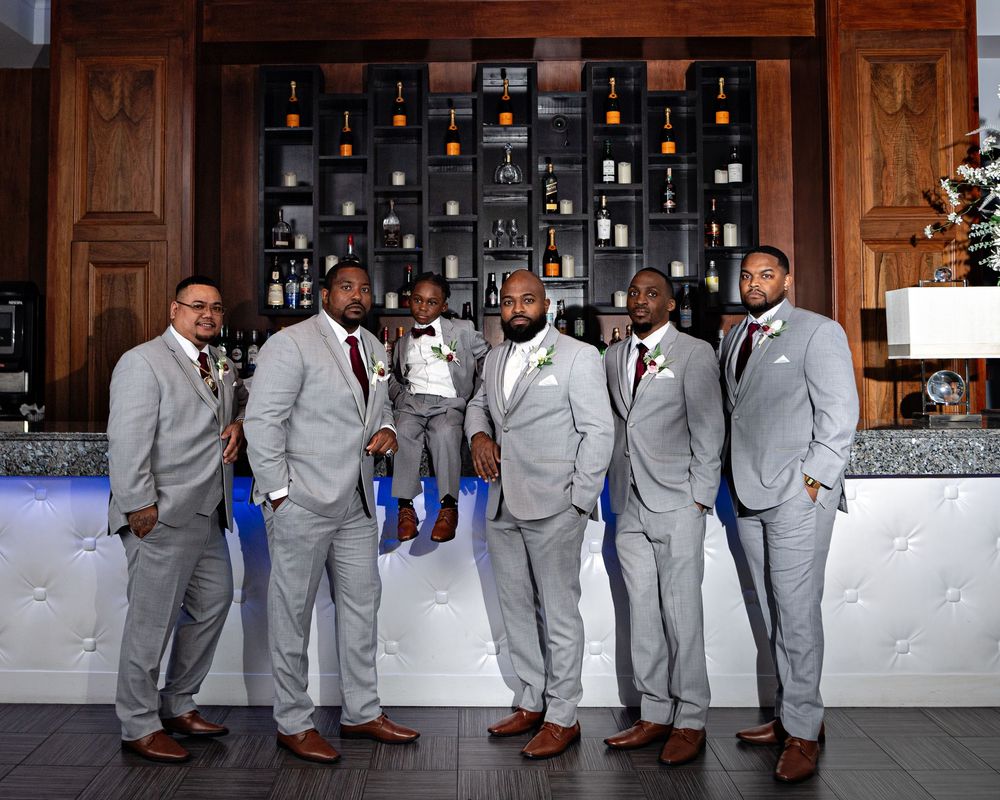 Groomsmen in light gray suits and brown shoes pose together in sequence at a wedding reception venue.