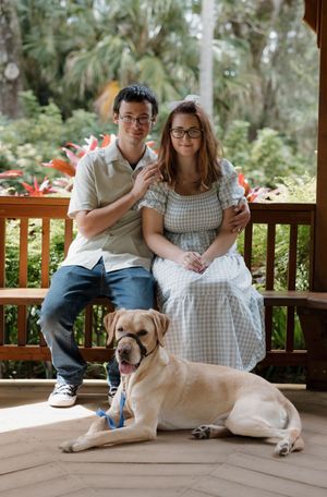 A couple sits on a wooden deck with their tan Labrador dog wearing a blue bandana.