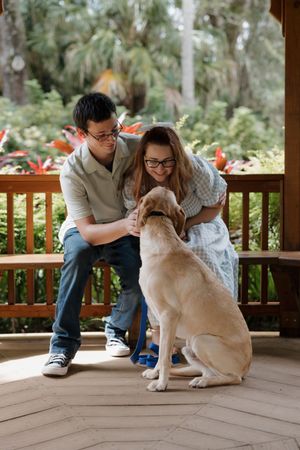 A loving couple pets their tan Labrador on a wooden deck surrounded by tropical foliage.