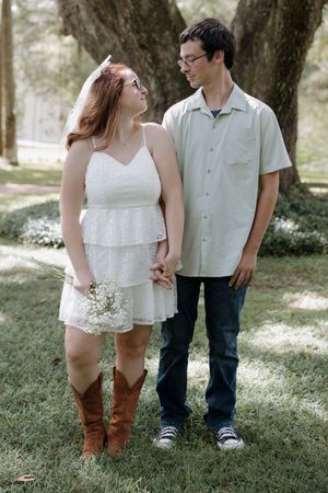 A couple stands together on grass, wearing contrasting casual and dressy outfits.