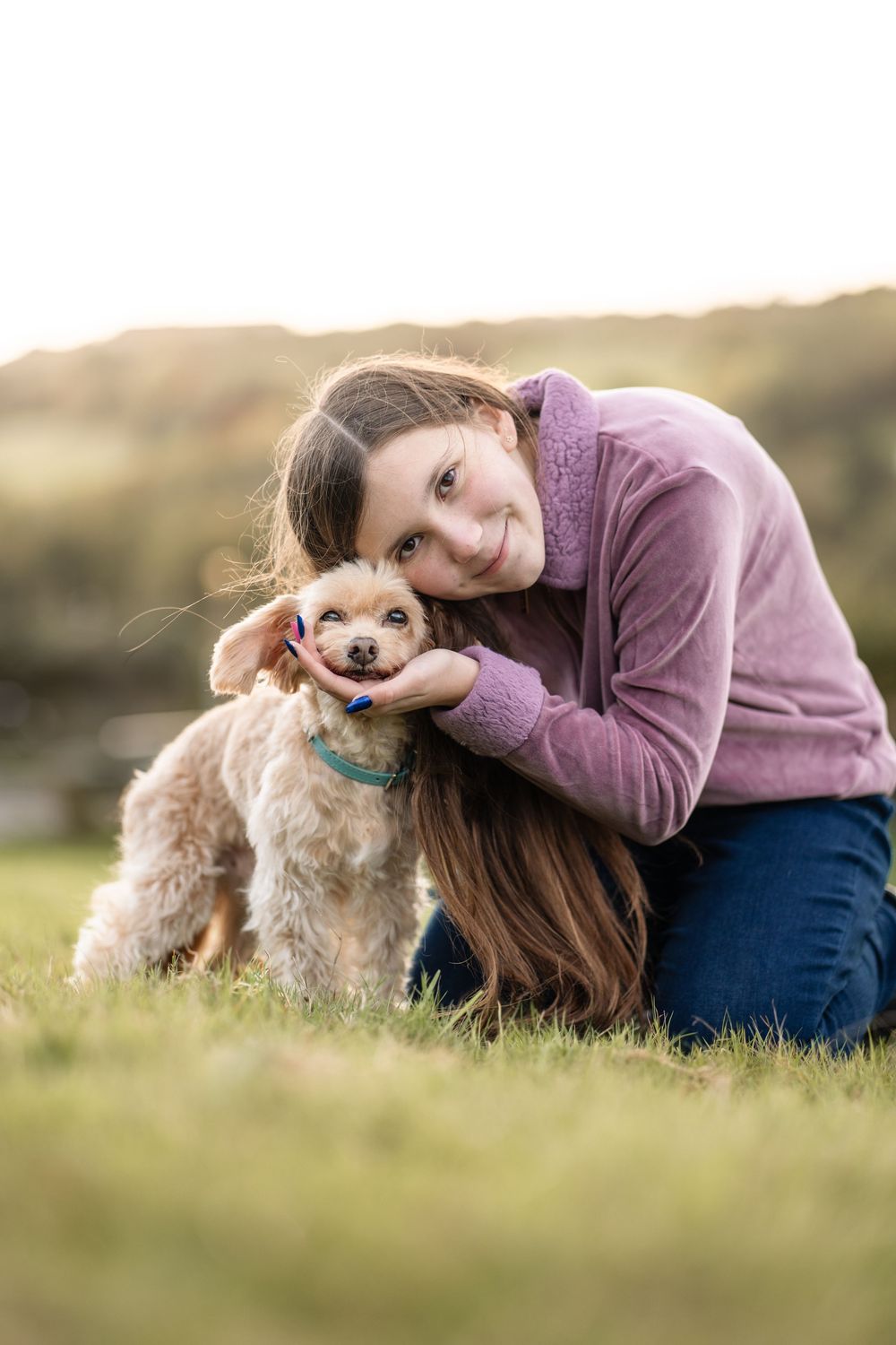 A person in a purple sweater cuddles with their small brown dog on grassy ground during golden hour.
