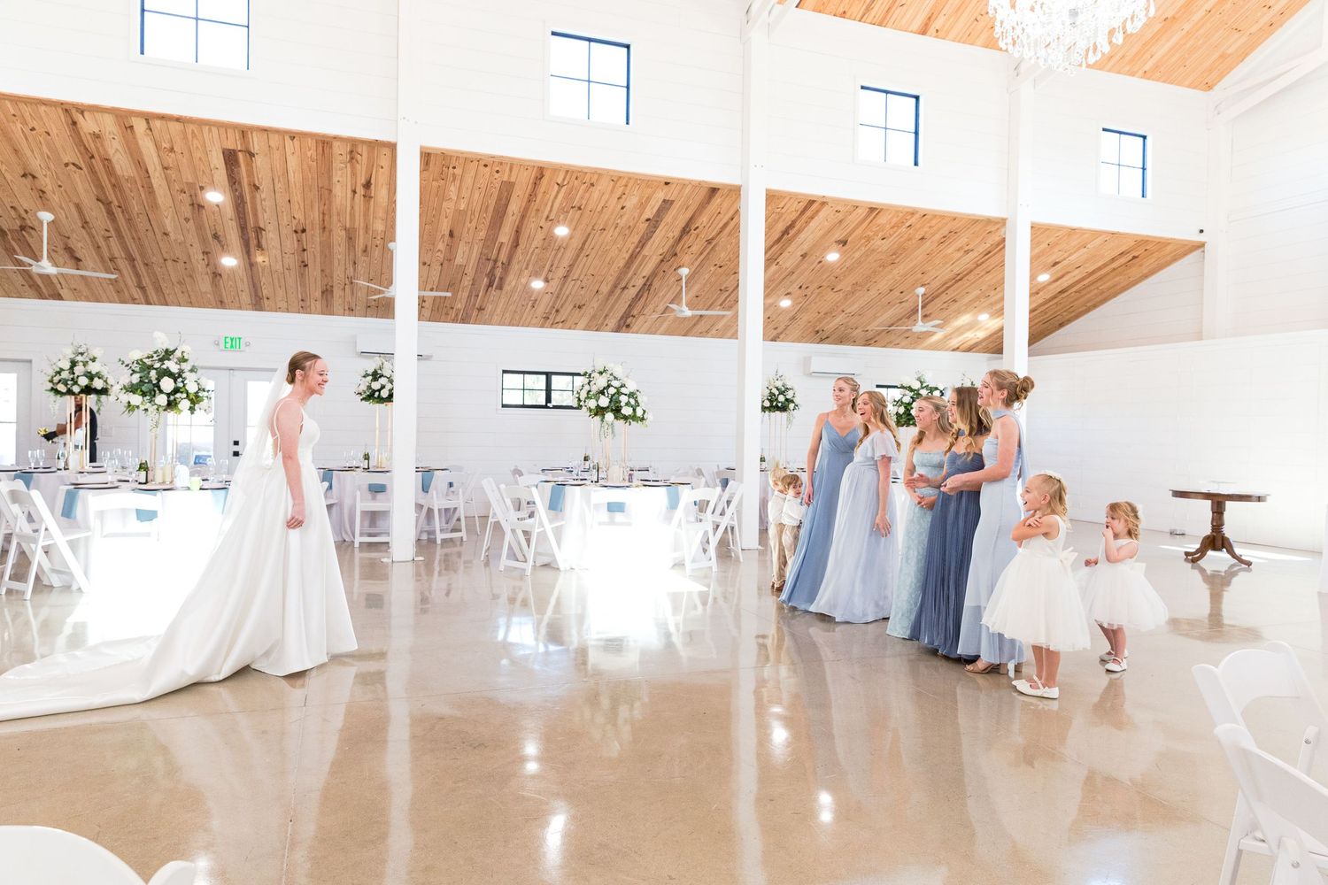 Bride in white gown shares first look with bridesmaids in blue dresses and flower girls in a bright white wedding venue.
