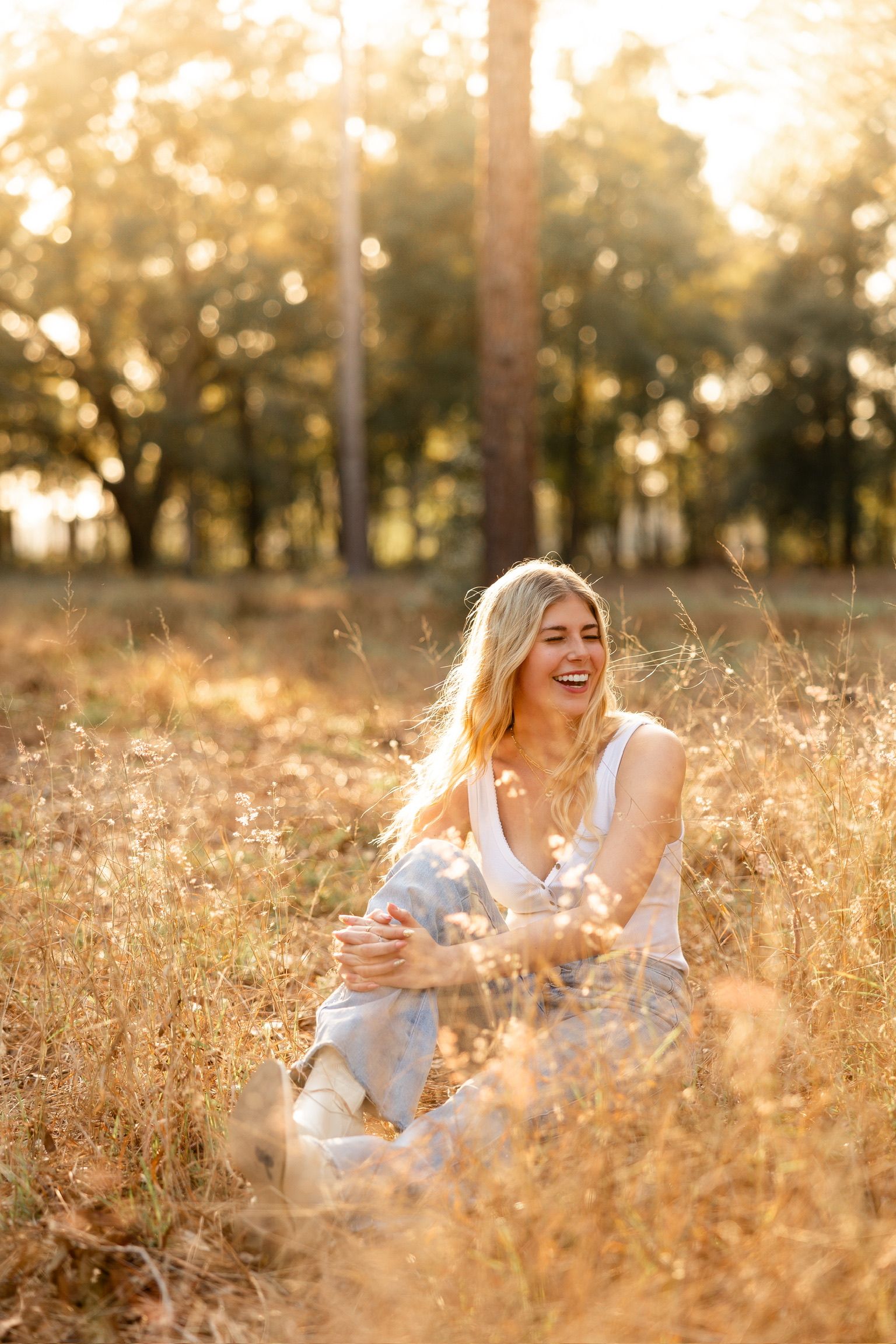 Woman in white top laughing while sitting in golden field during sunset.