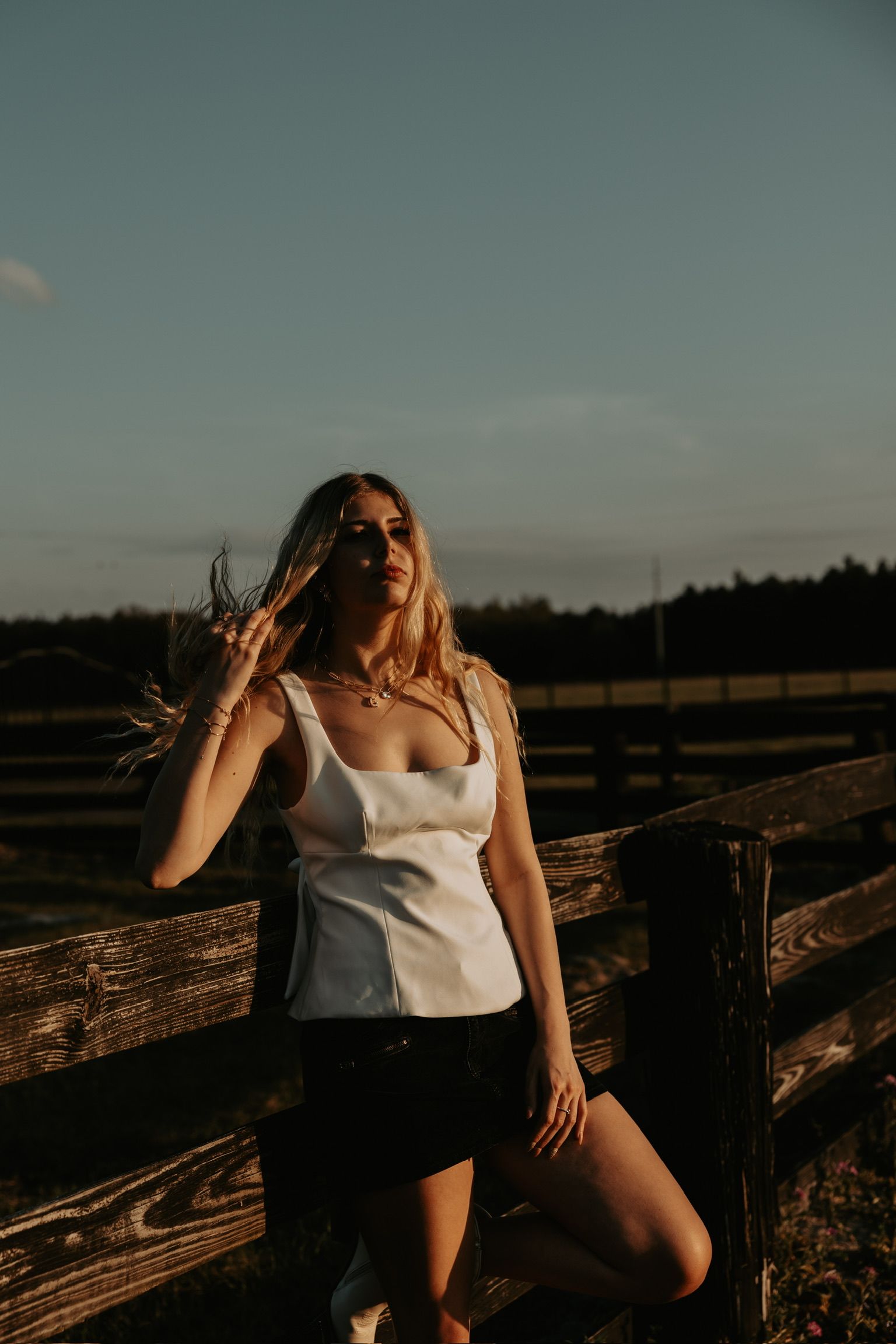 Woman in white top posing on rustic wooden fence at dusk.
