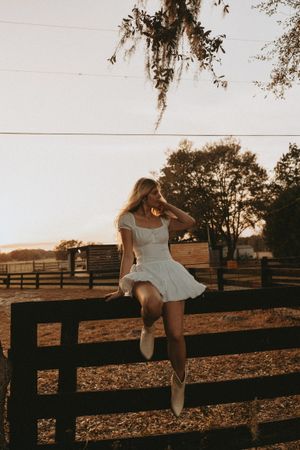A white sundress contrasts against a dark wooden fence during golden hour in a countryside setting.