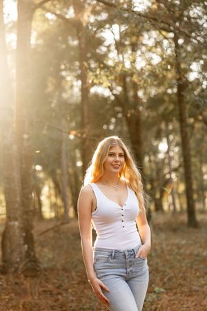 Golden sunlight filters through trees as a person in a white tank top and jeans poses in a wooded setting.