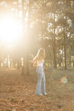 Person in light denim jeans stands in a sunlit autumn forest with golden light filtering through trees.