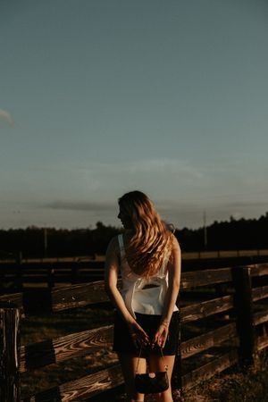 Person in casual attire leans against rustic wooden fence at sunset in countryside setting.