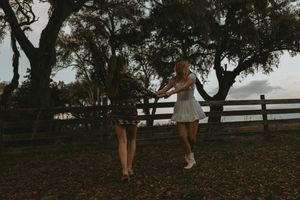 Two people in white dresses dance together under moody trees at dusk with a wooden fence in the background.