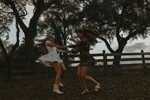 Two dancers in flowing white dresses leap gracefully beneath dark trees with a rustic fence behind them.