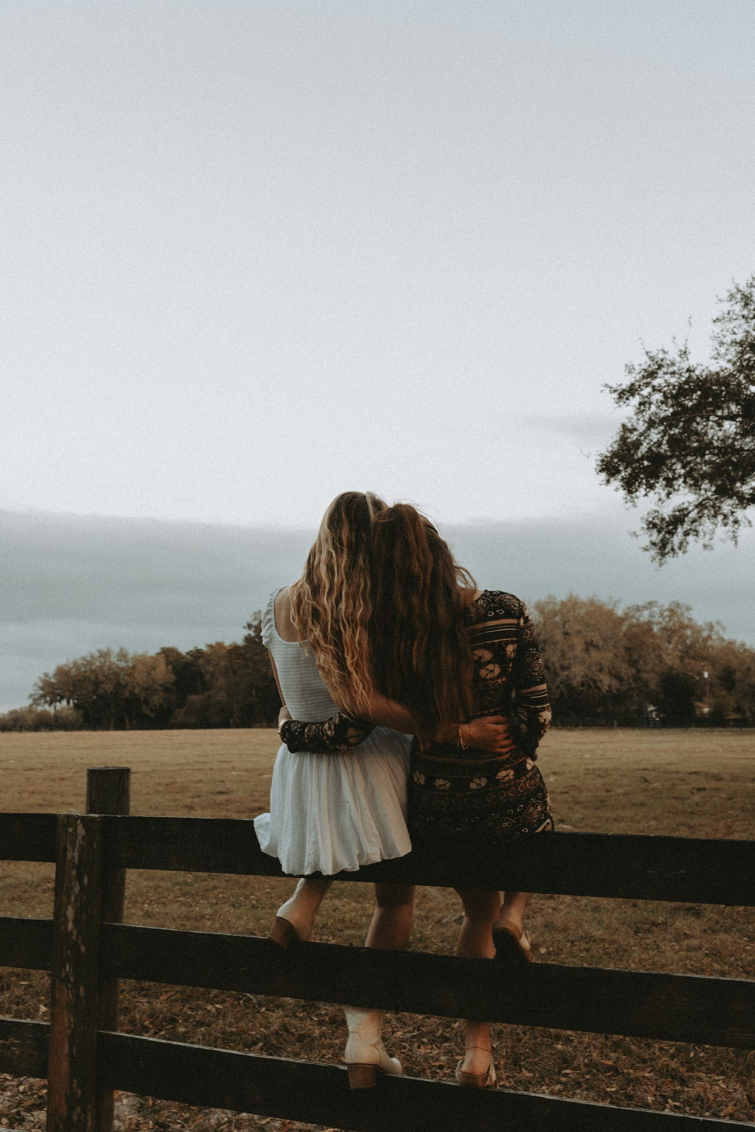 Person in white dress sitting on rustic wooden fence at sunset in countryside setting.