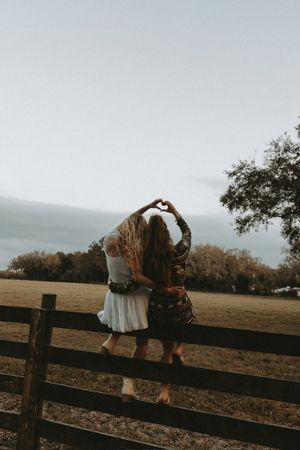 Two people sit on a wooden fence making a heart shape with their arms against a moody autumn landscape.