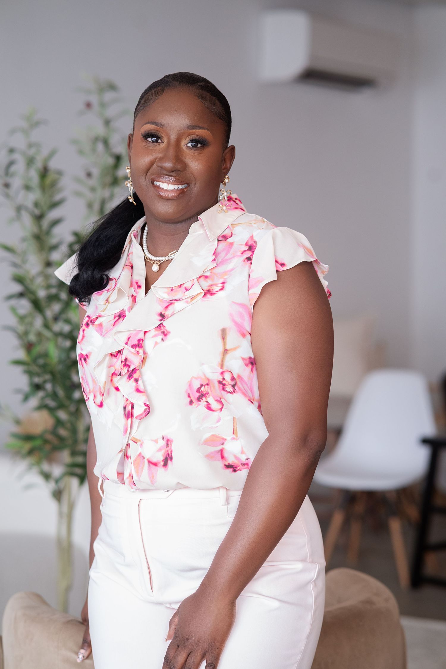 A person wearing a white and pink floral blouse with white pants stands smiling in a modern interior setting.