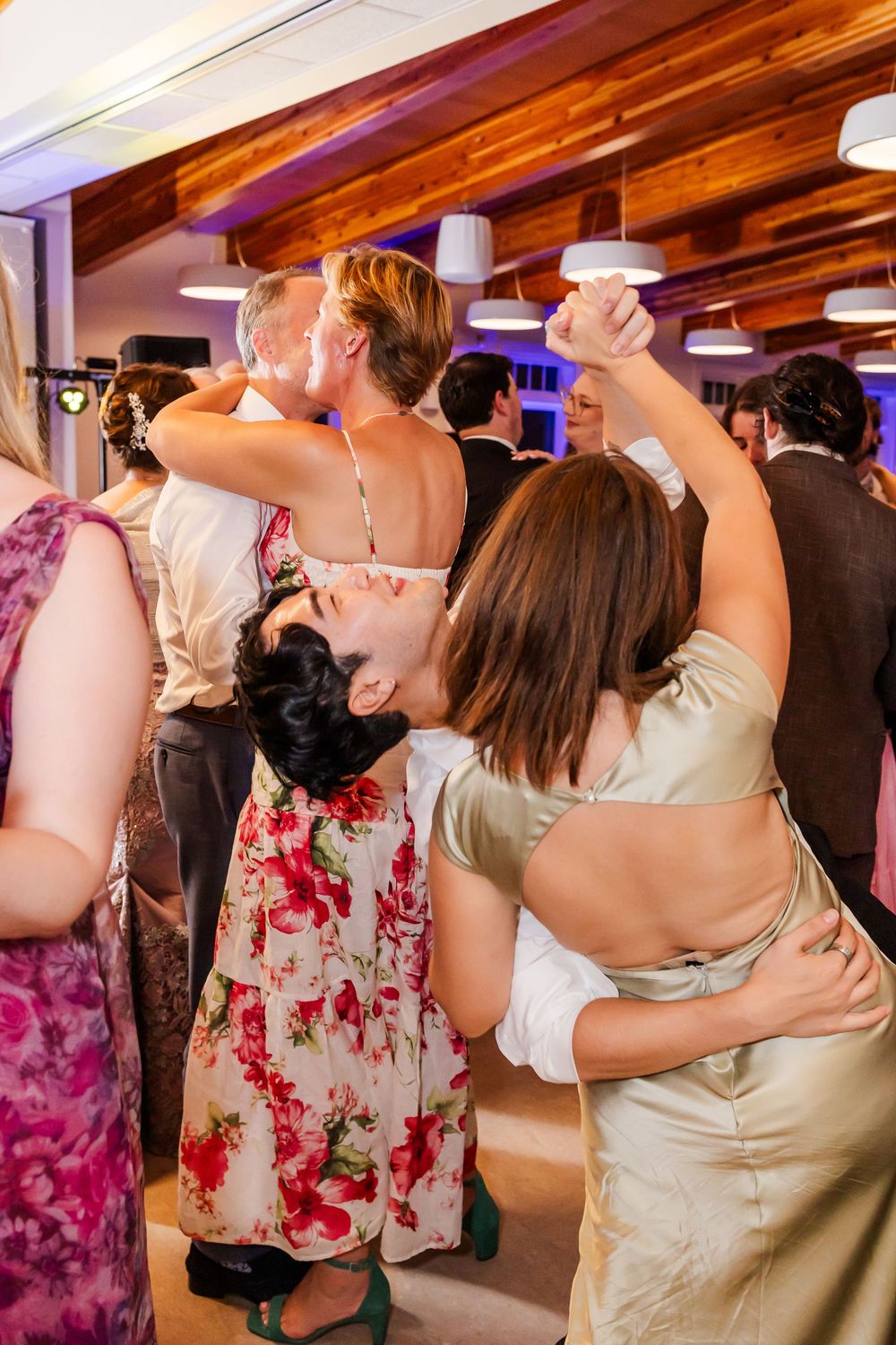 Wedding guests dance and embrace on a crowded dance floor illuminated by colorful party lights.