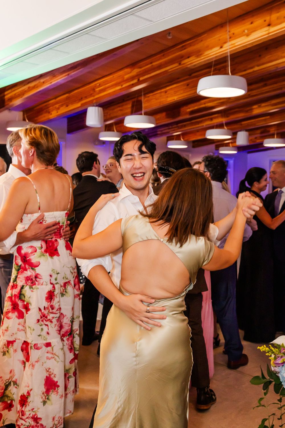 Wedding guests dance together under wooden beams and pendant lights at an indoor reception celebration.