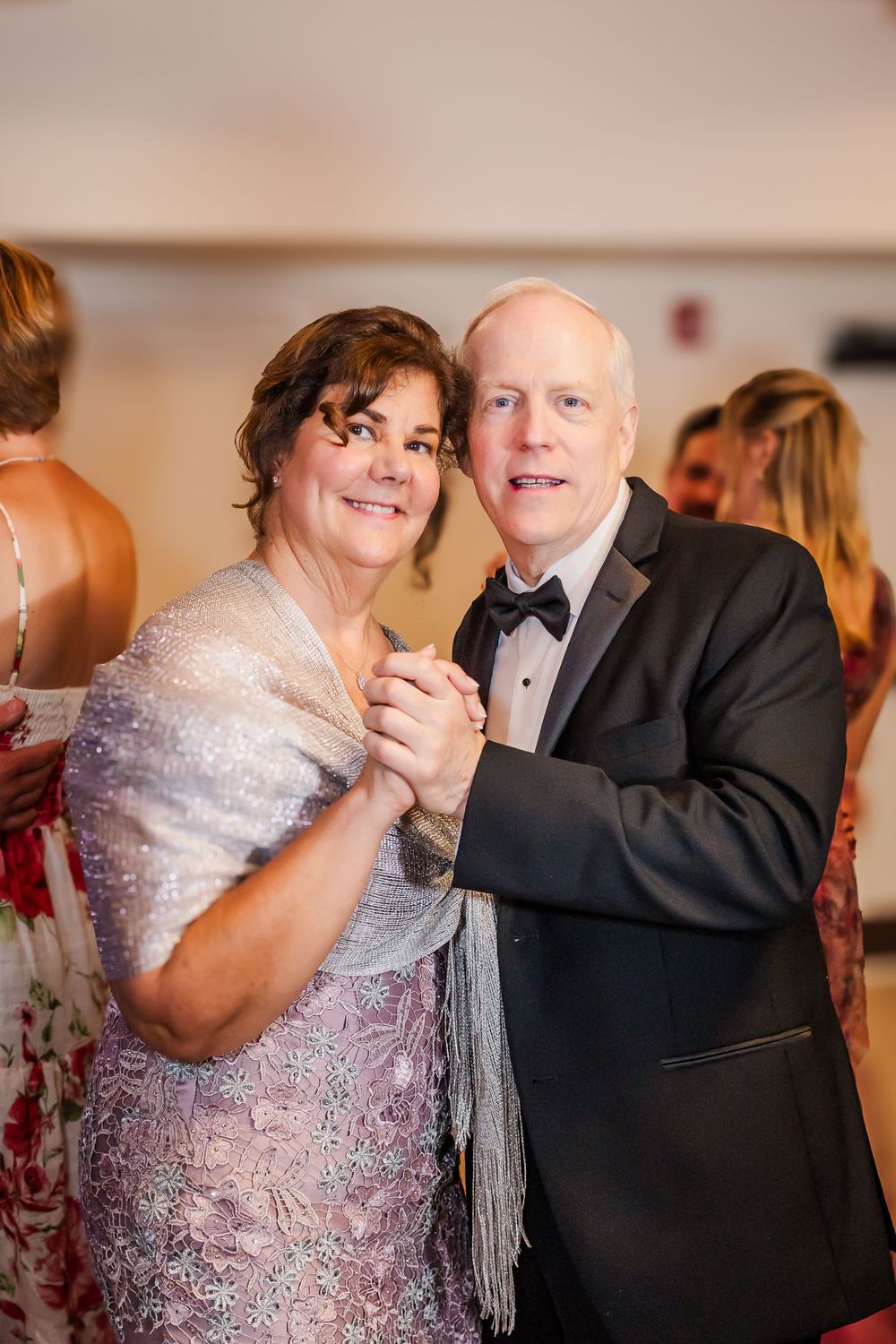 An elegant couple dances together at a formal event, with the woman wearing a sparkly dress and silver shawl.