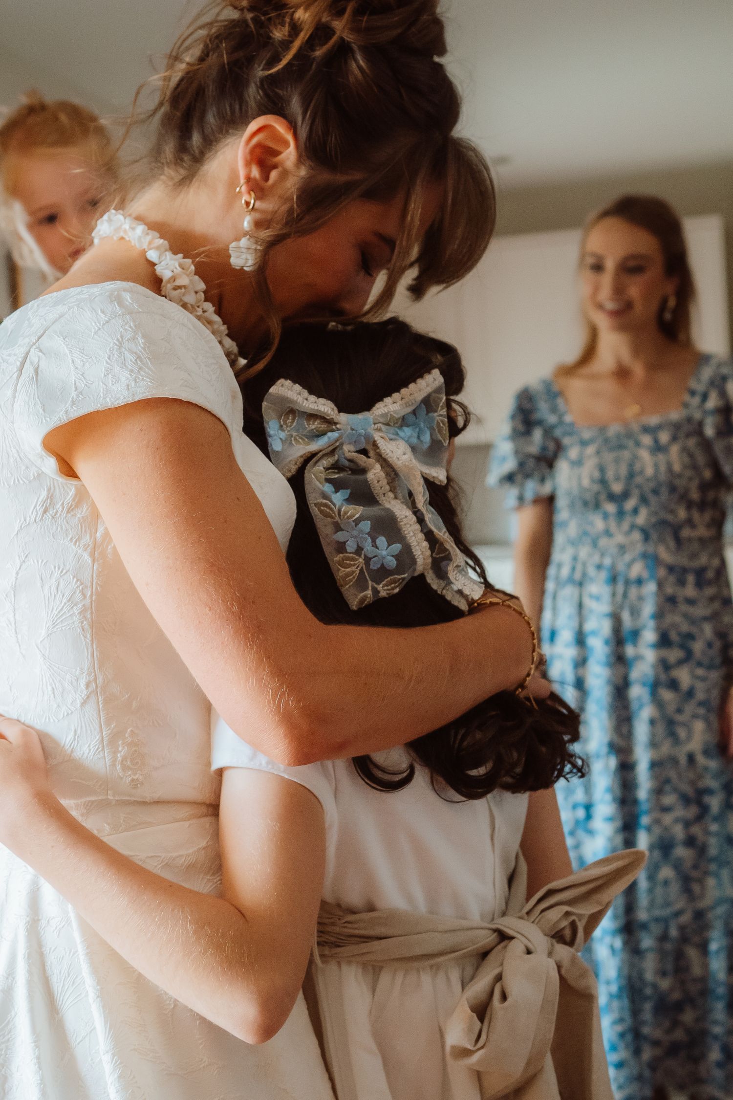 A bride in a white lace dress embraces someone while bridesmaids in floral blue dresses stand nearby.