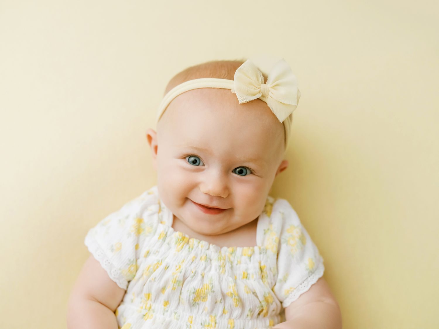 A smiling baby wearing a floral dress and cream bow headband against a beige background.