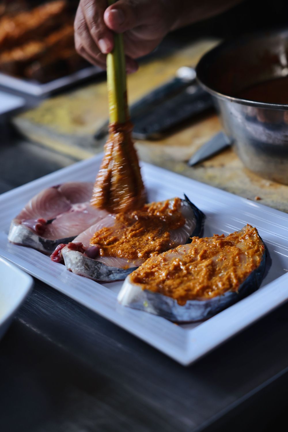 Orange sauce being spread on raw meat on a white plate in a dark setting.