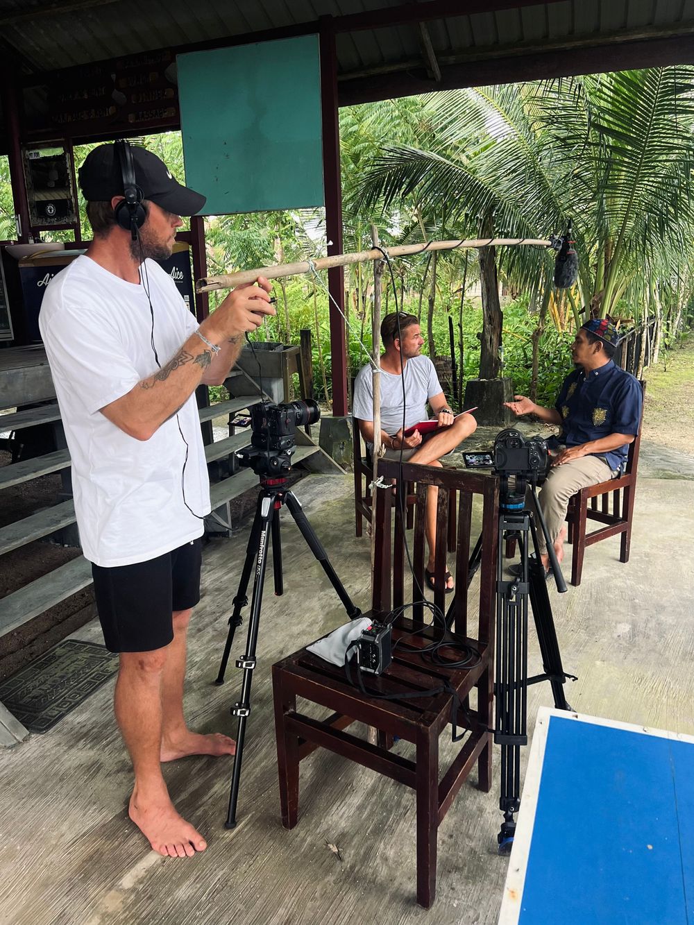 Film crew setting up camera equipment on wooden stools in an outdoor tropical setting.