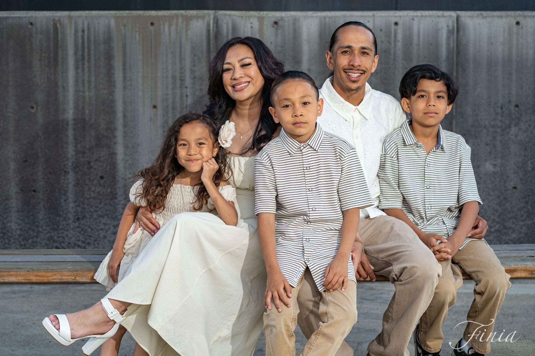 Family wearing coordinated white and beige outfits sits together on steps for a professional outdoor photo shoot.