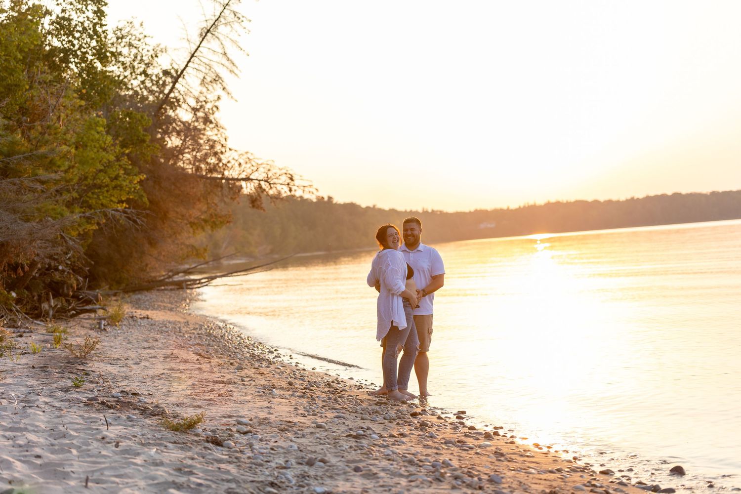 A couple embraces on a sandy beach at golden sunset along a tranquil rivershore.