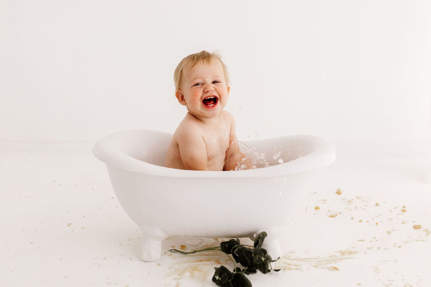 Baby splashing and playing during bath time photo session in white tub.