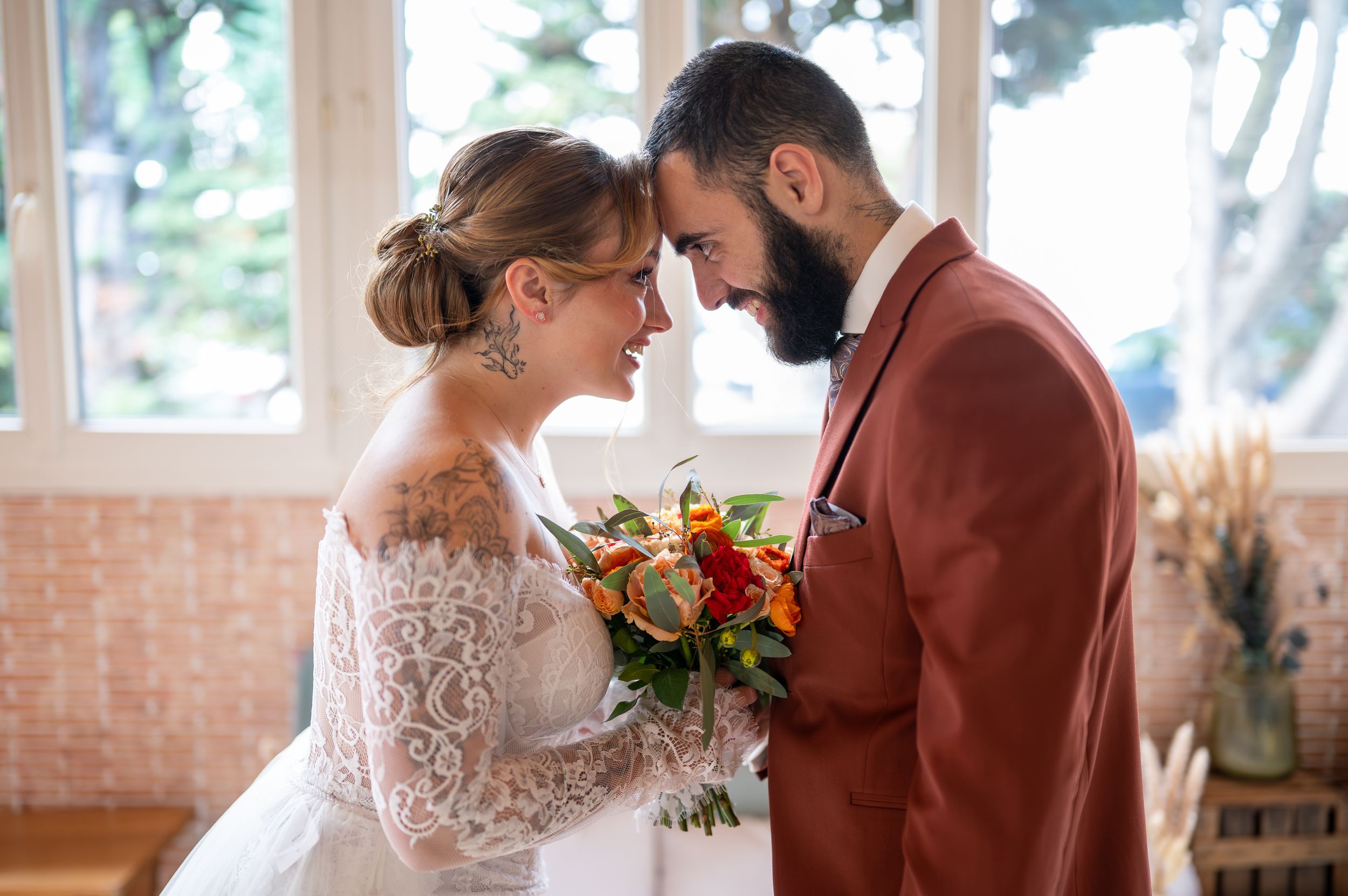Un couple en tenue de mariage partage un moment intime dans une pièce ensoleillée avec des murs et des fenêtres en briques rustiques.