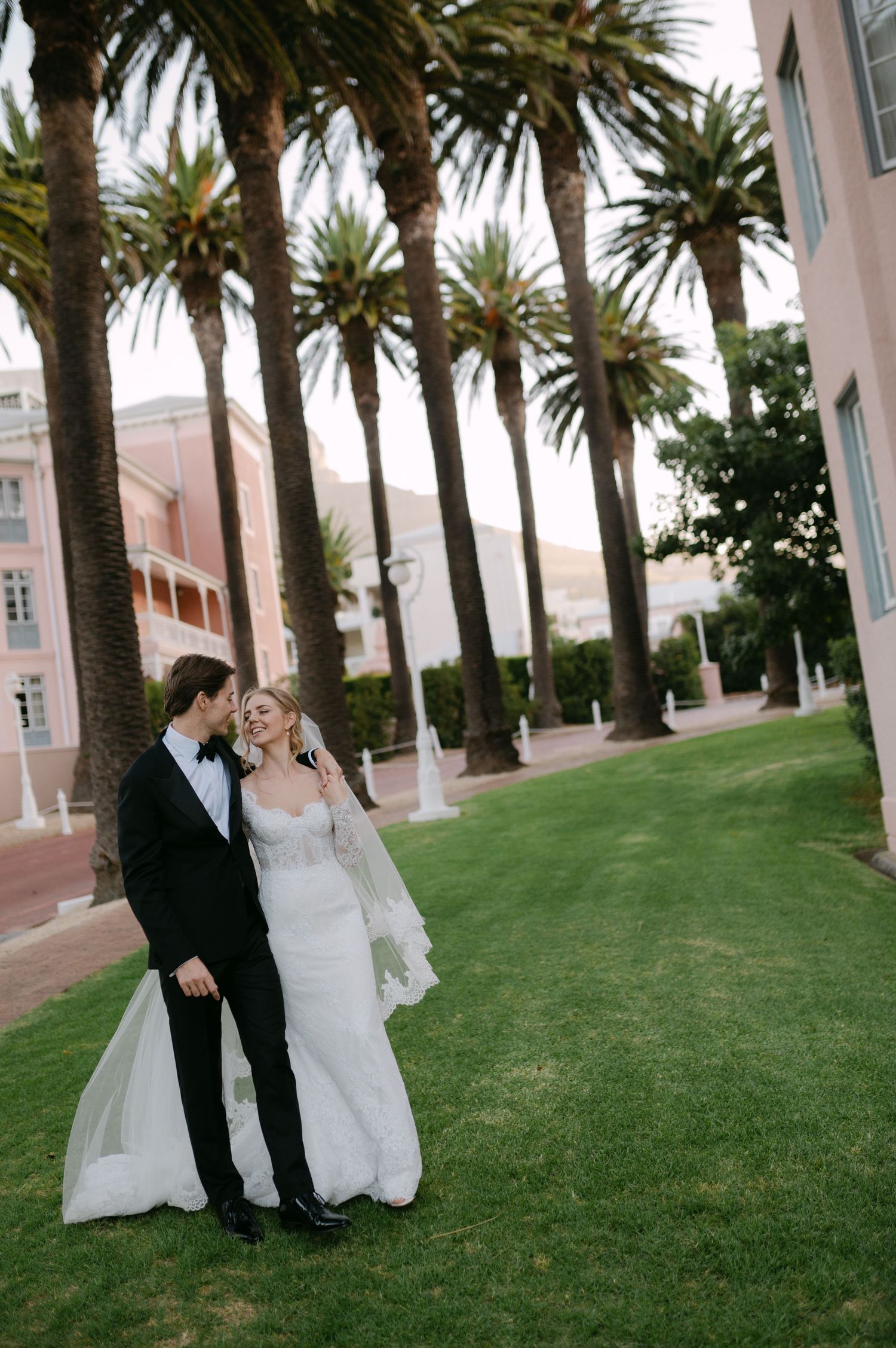 A romantic wedding couple poses along a palm tree-lined path on a manicured lawn at a luxury resort.