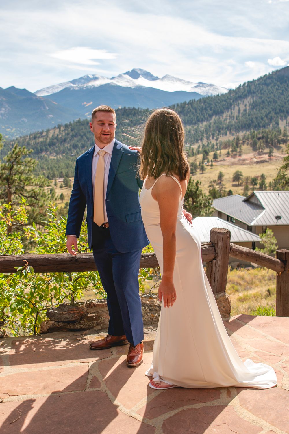 A couple in formal wedding attire stands on a mountain terrace overlooking snow-capped peaks and evergreen forests.