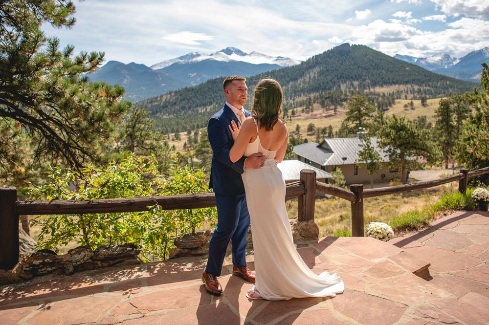 A couple stands at a scenic mountain overlook with snow-capped peaks and evergreen forests in the background.