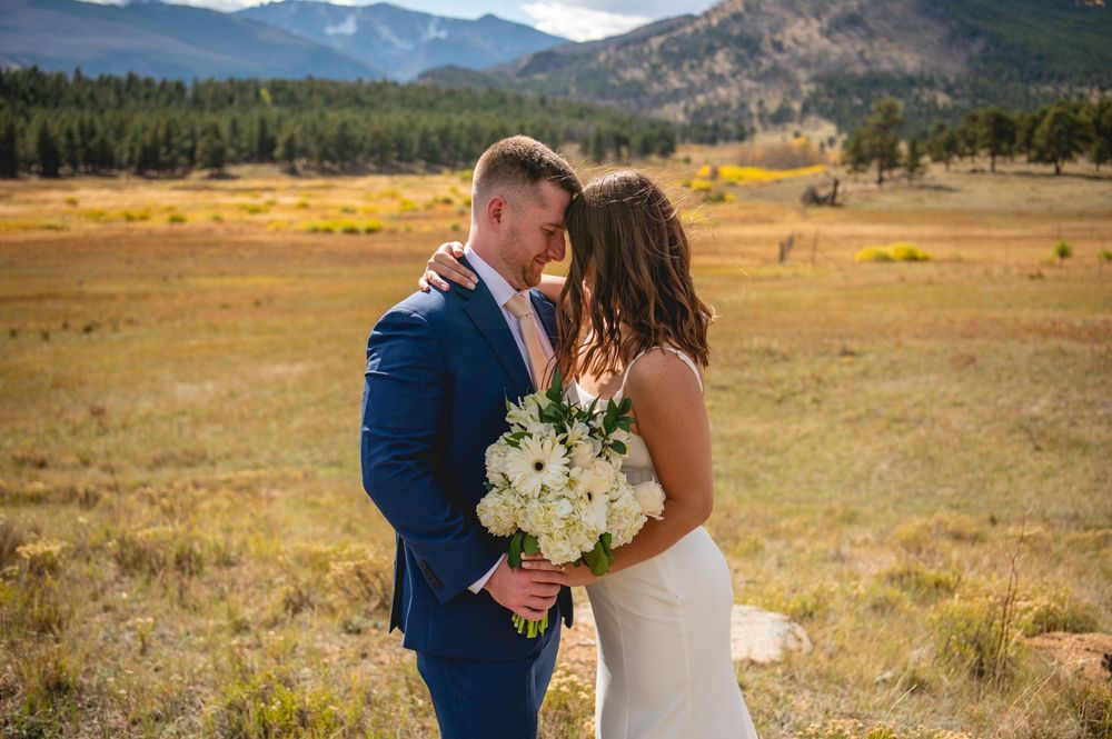 A couple shares a romantic moment in a scenic mountain meadow with golden grass and evergreen forests.